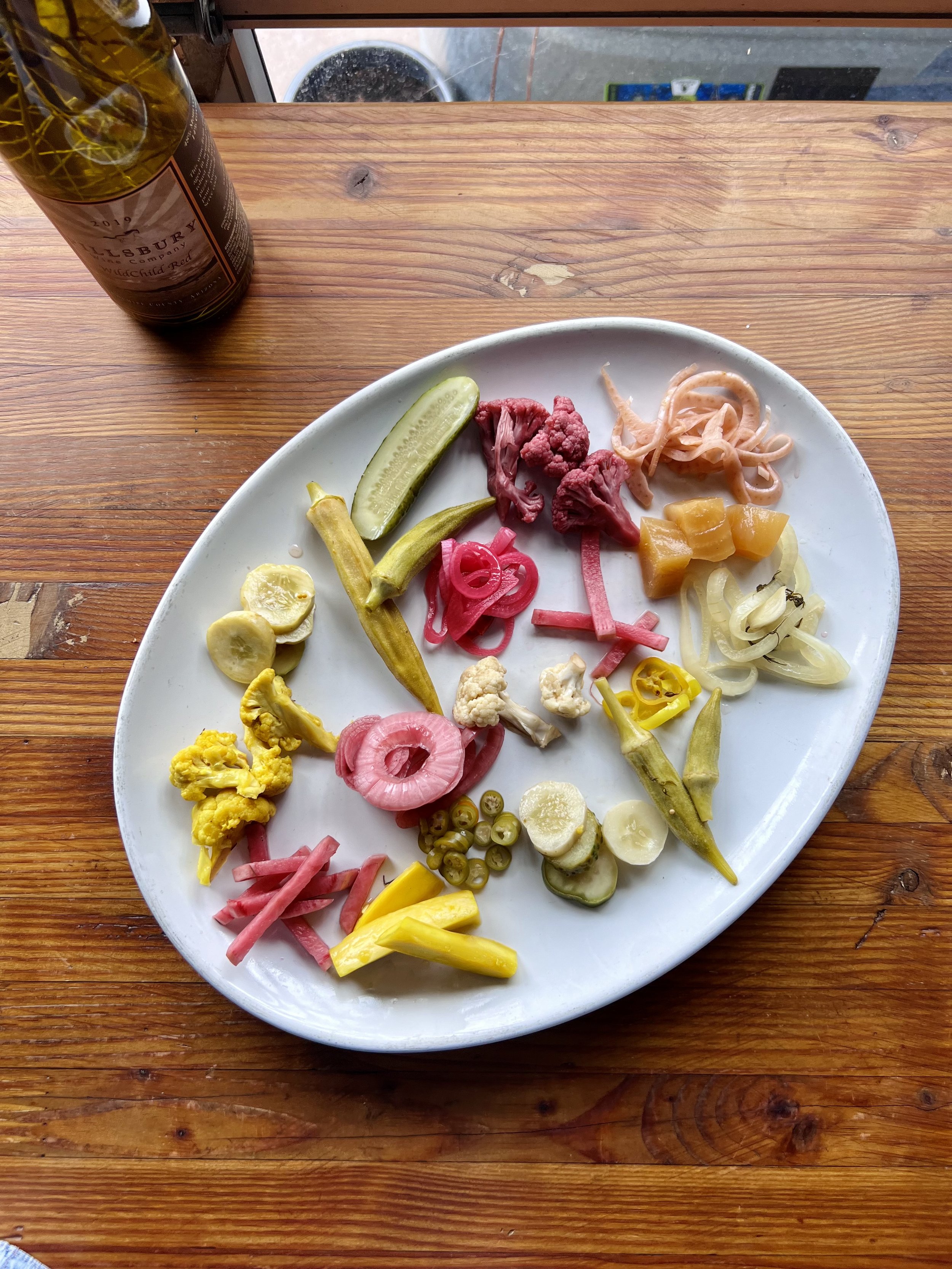 A white oval plate with assorted pickled vegetables including cucumbers, okra, cauliflower, radishes, onions, peppers, and squash on a wooden table.