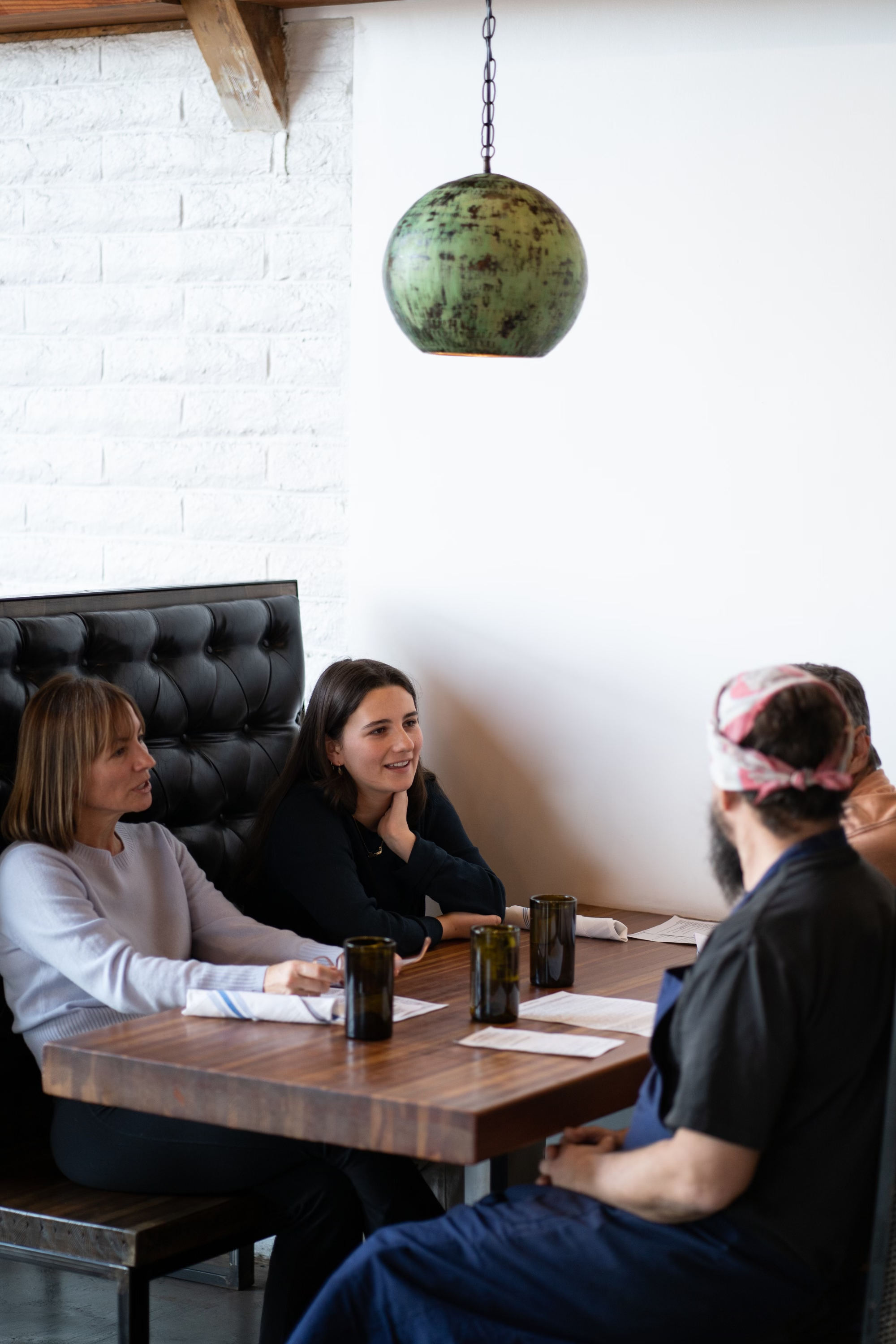 Group of four people sitting at a dining table in a restaurant, engaged in conversation, with a black leather backrest, white brick wall, and a green pendant light hanging above.