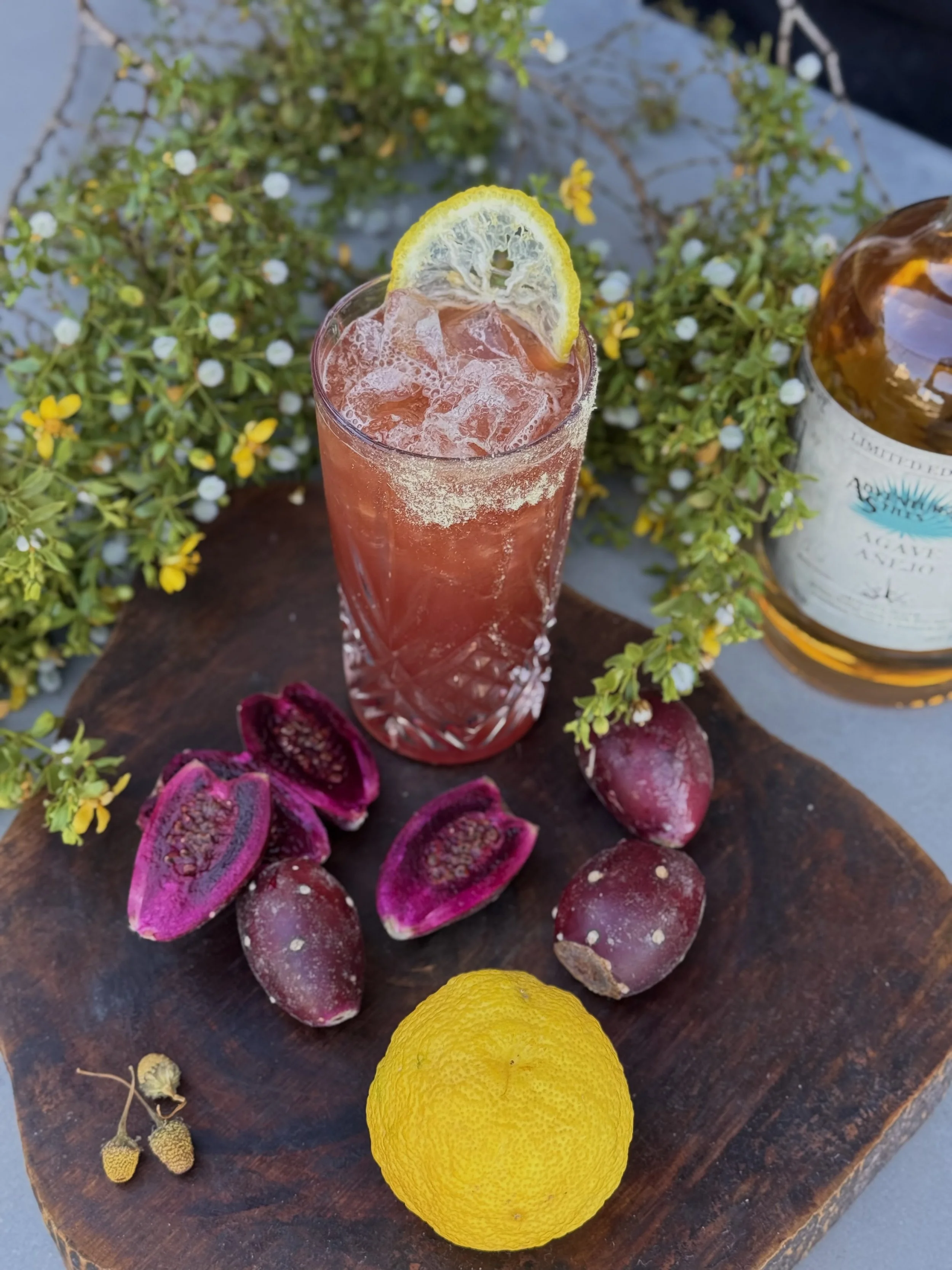 A glass of pink cocktail garnished with a lemon slice, surrounded by pink and purple sprouted fruits, a yellow lemon, greenery, and a bottle of alcohol on a wooden surface.