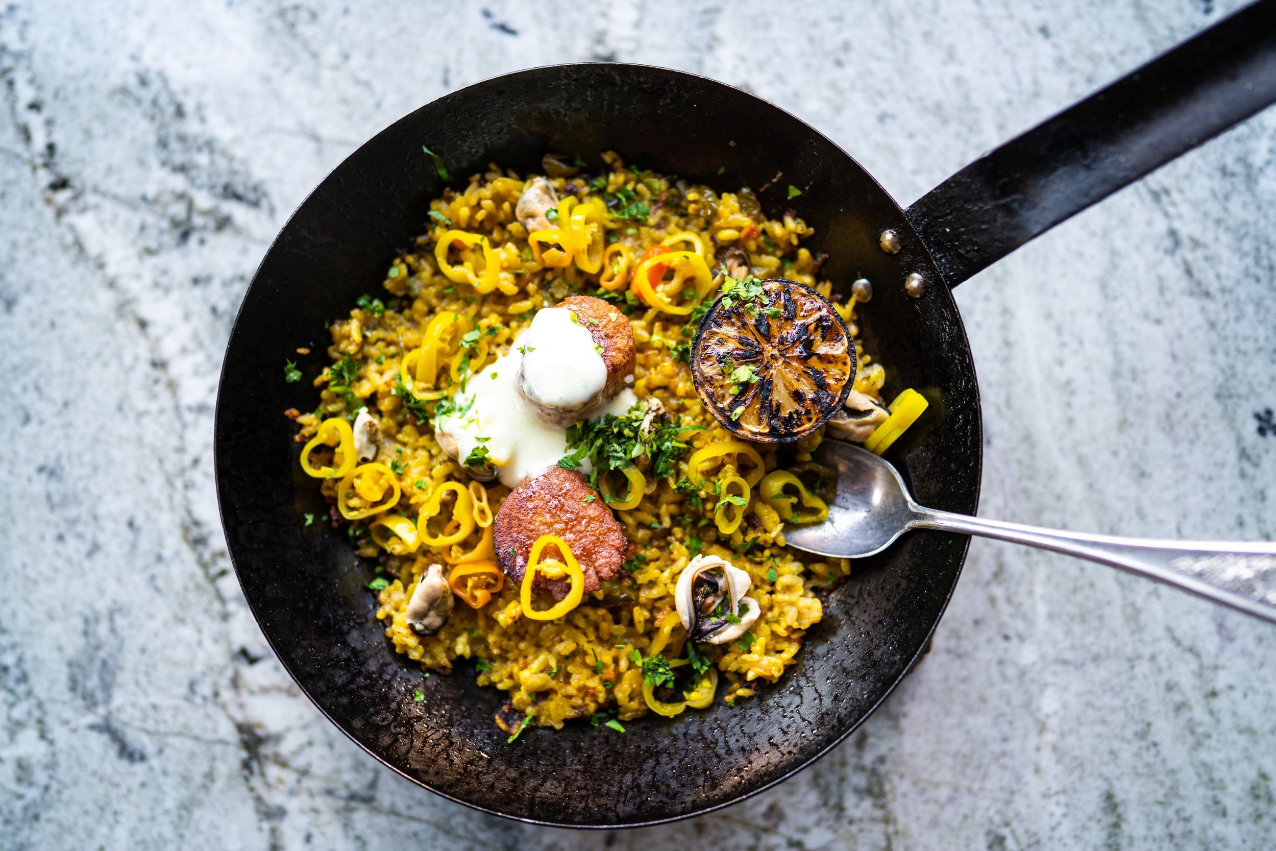 Black skillet filled with yellow rice, sliced yellow peppers, grilled lemon, sausage slices, dollops of white sauce, mushrooms, chopped green herbs, and a silver spoon, on a textured light surface.