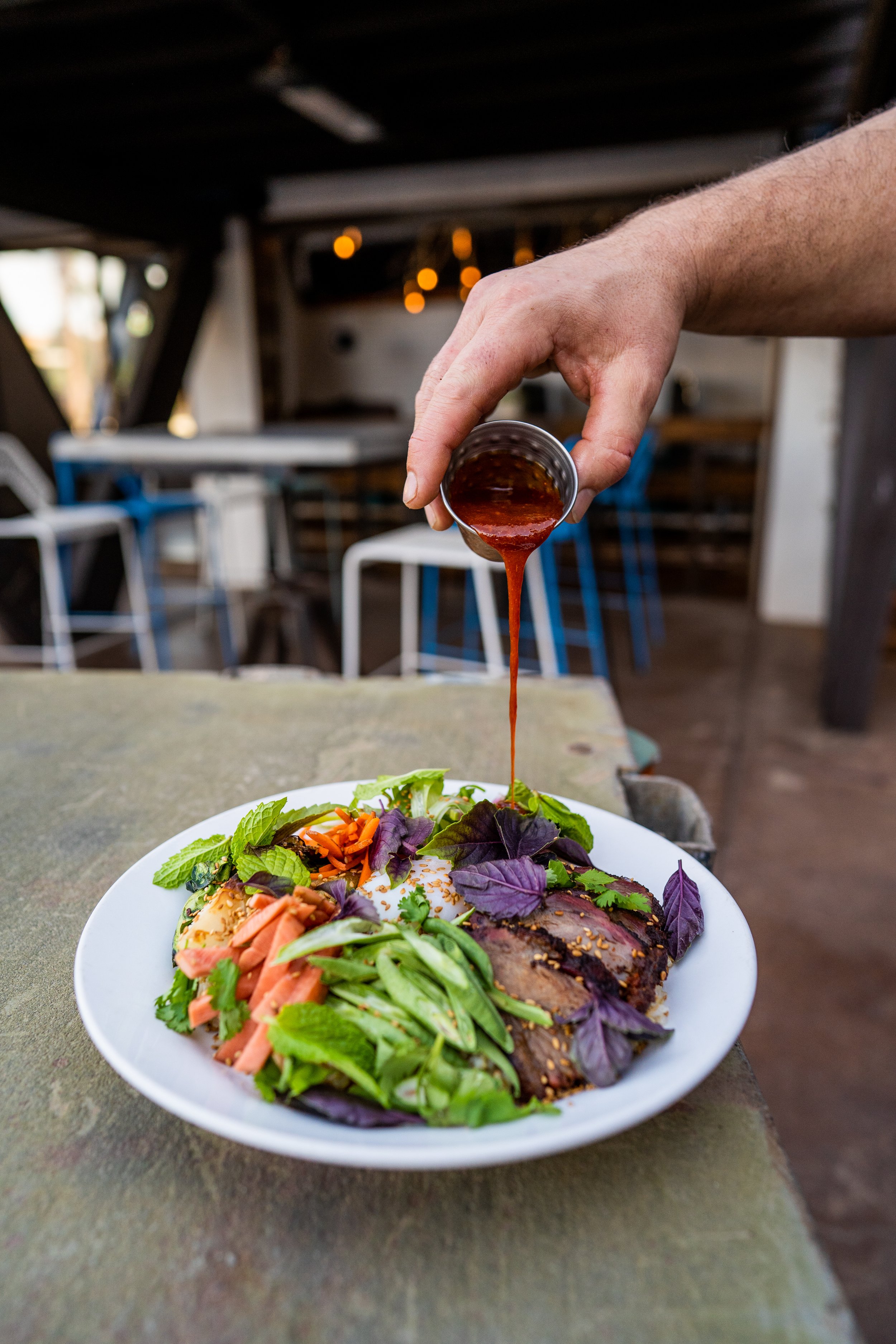 Person pouring sauce onto a plate of food that includes sliced meat, vegetables, and a soft-boiled egg, in a rustic restaurant setting.