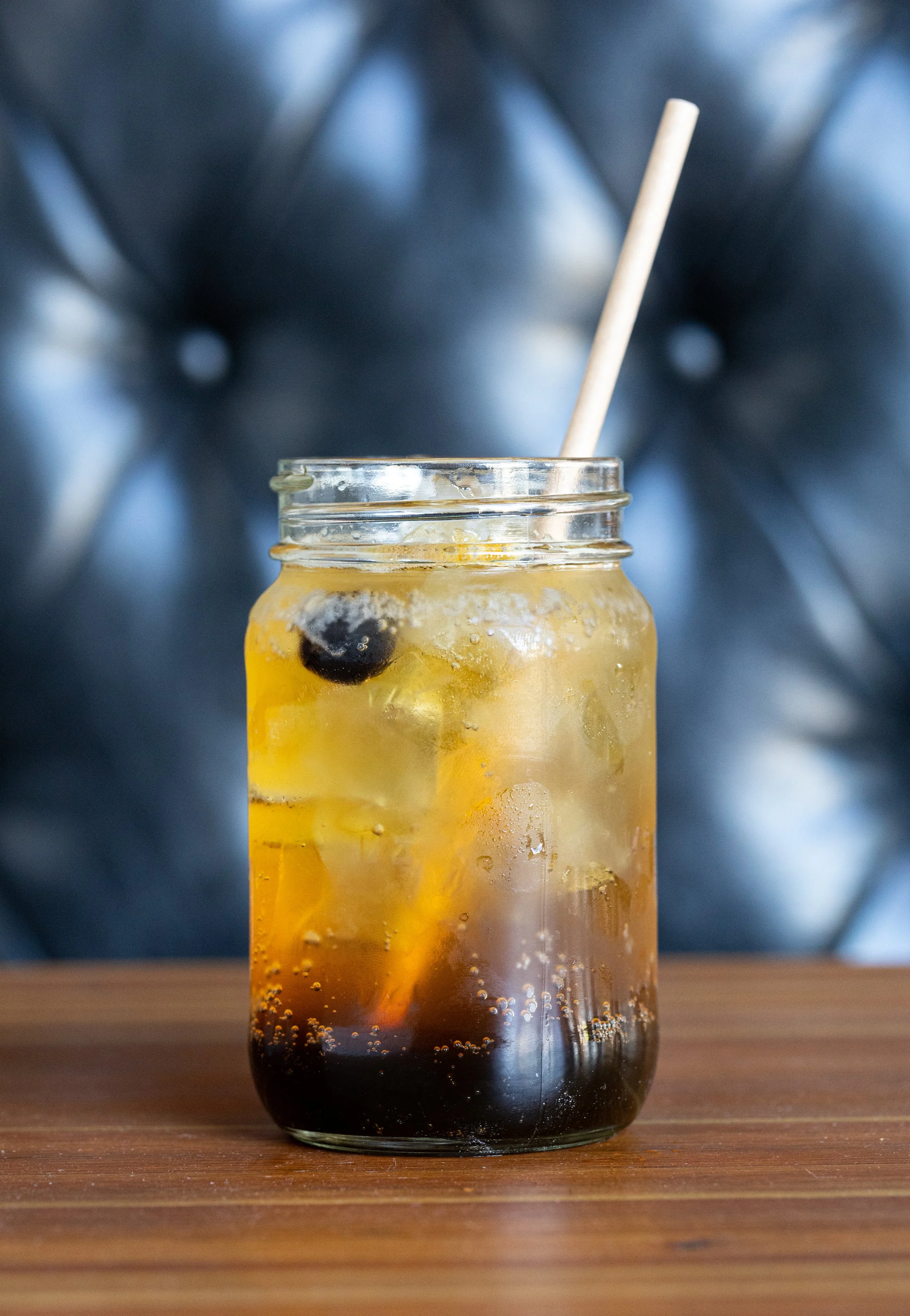 A glass jar filled with iced tea, ice cubes, and a straw, sitting on a wooden surface.