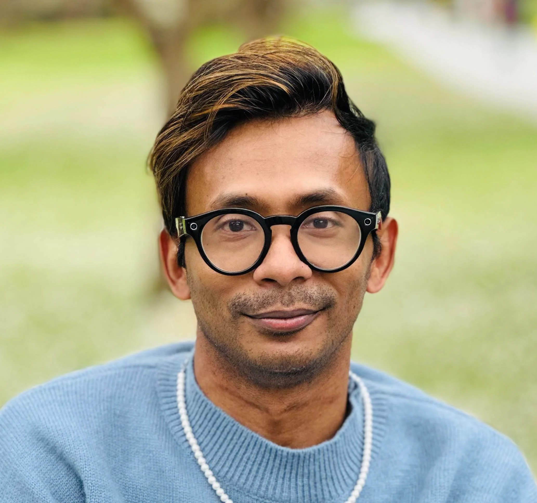 Headshot of Ather Sharif, a brown man with black and brown hair smiling at the camera, wearing eyeglasses, a light blue sweater, and a pearl necklace. The background is blurred.