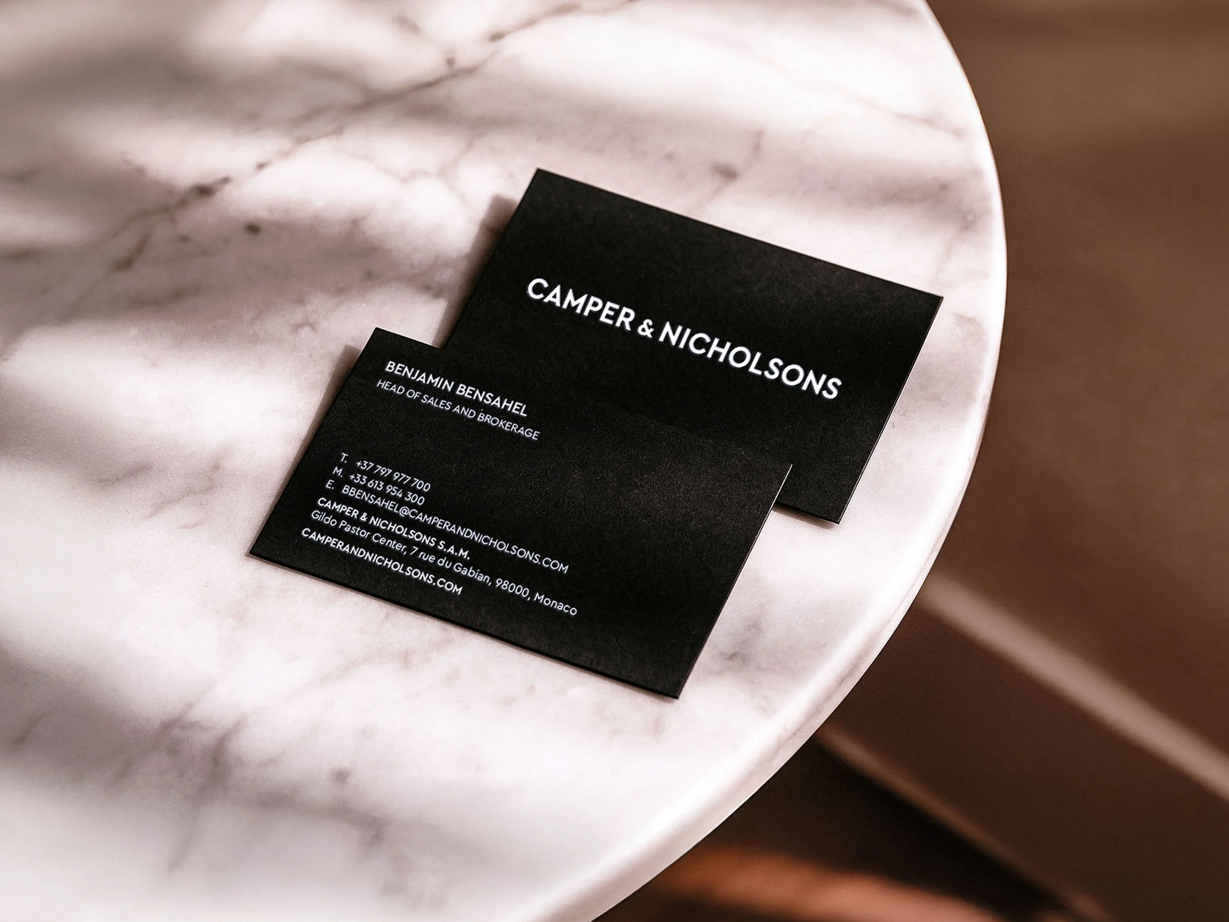 The Camper & Nicholsons business cards on a marble table. One card shows the front, with the logotype sitting in white in the centre. The other card shows a person's details. Both cards are black and all text is white