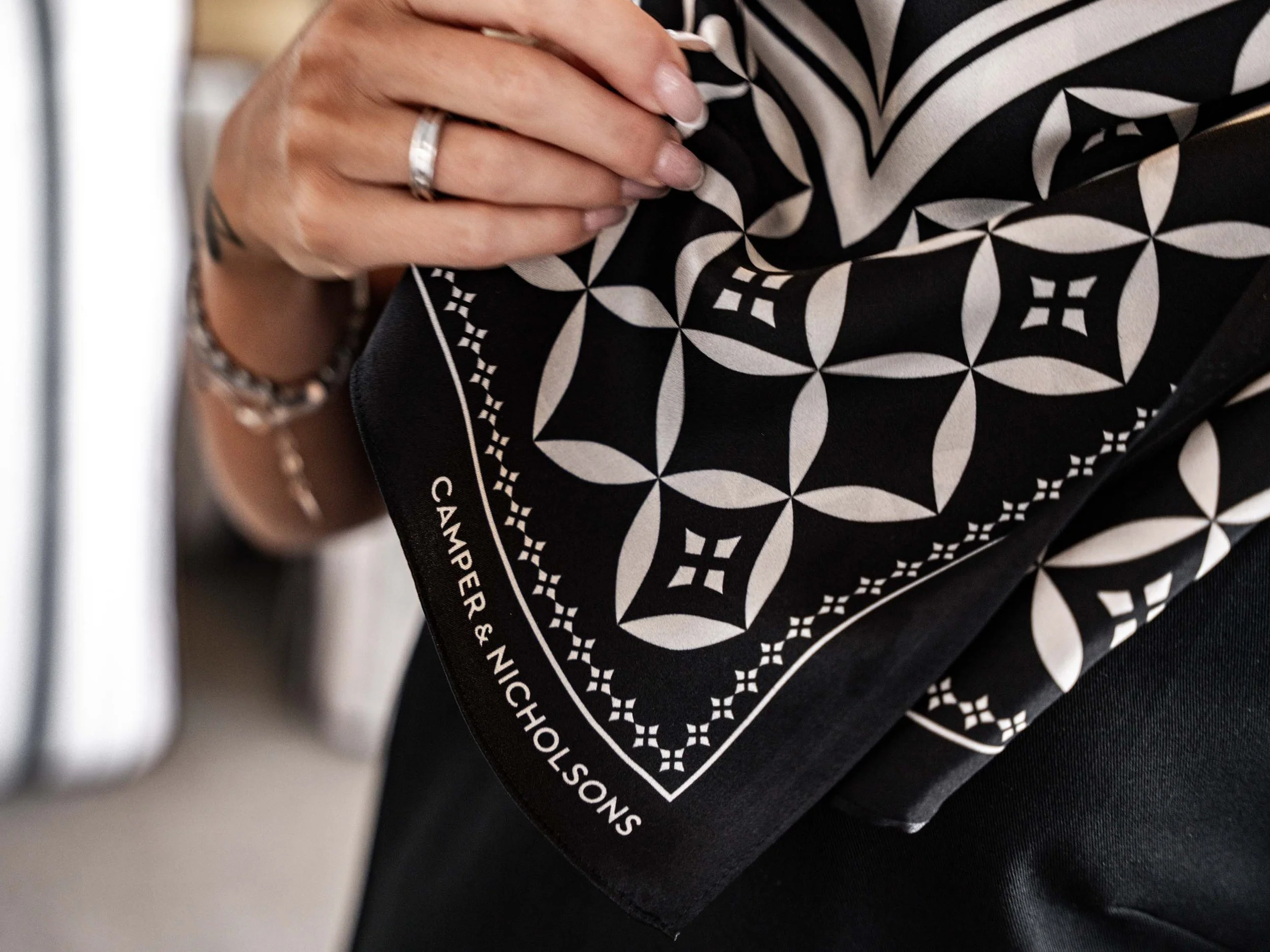 Image of a woman's hands holding the Camper & Nicholsons silk scarf. The scarf has the C&N pattern and the logotype printed on it. It is black and cream