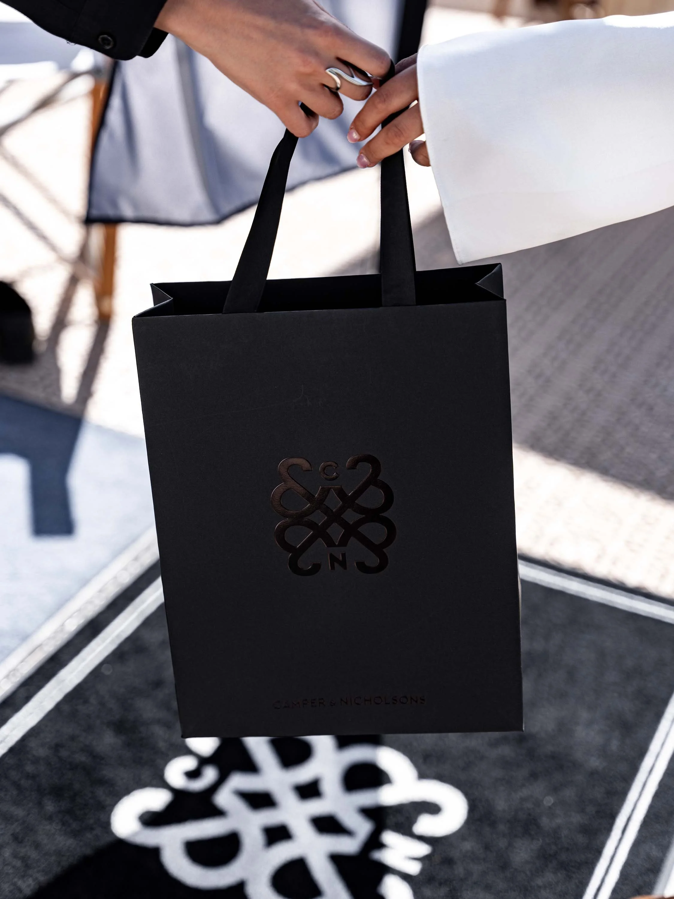 An image of two hand passing a C&N black carrier bag. The bag has the logo and emblem foiled in shiny black