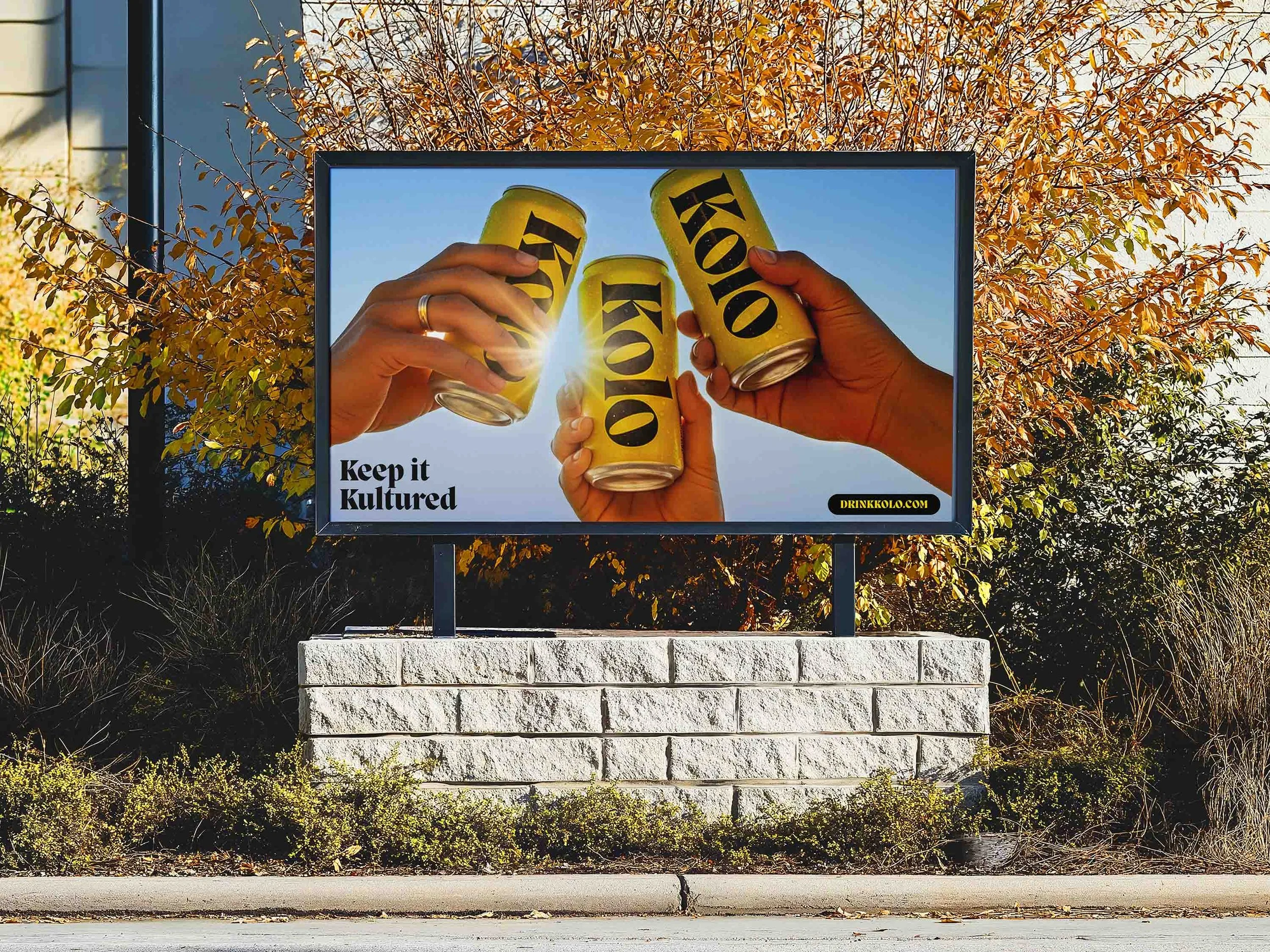 Image of a small billboard with a Kolo poster inside. The poster shows 3 hands holding the yellow Kolo cans. Small text in the bottom corner says 'Keep it Kultured"