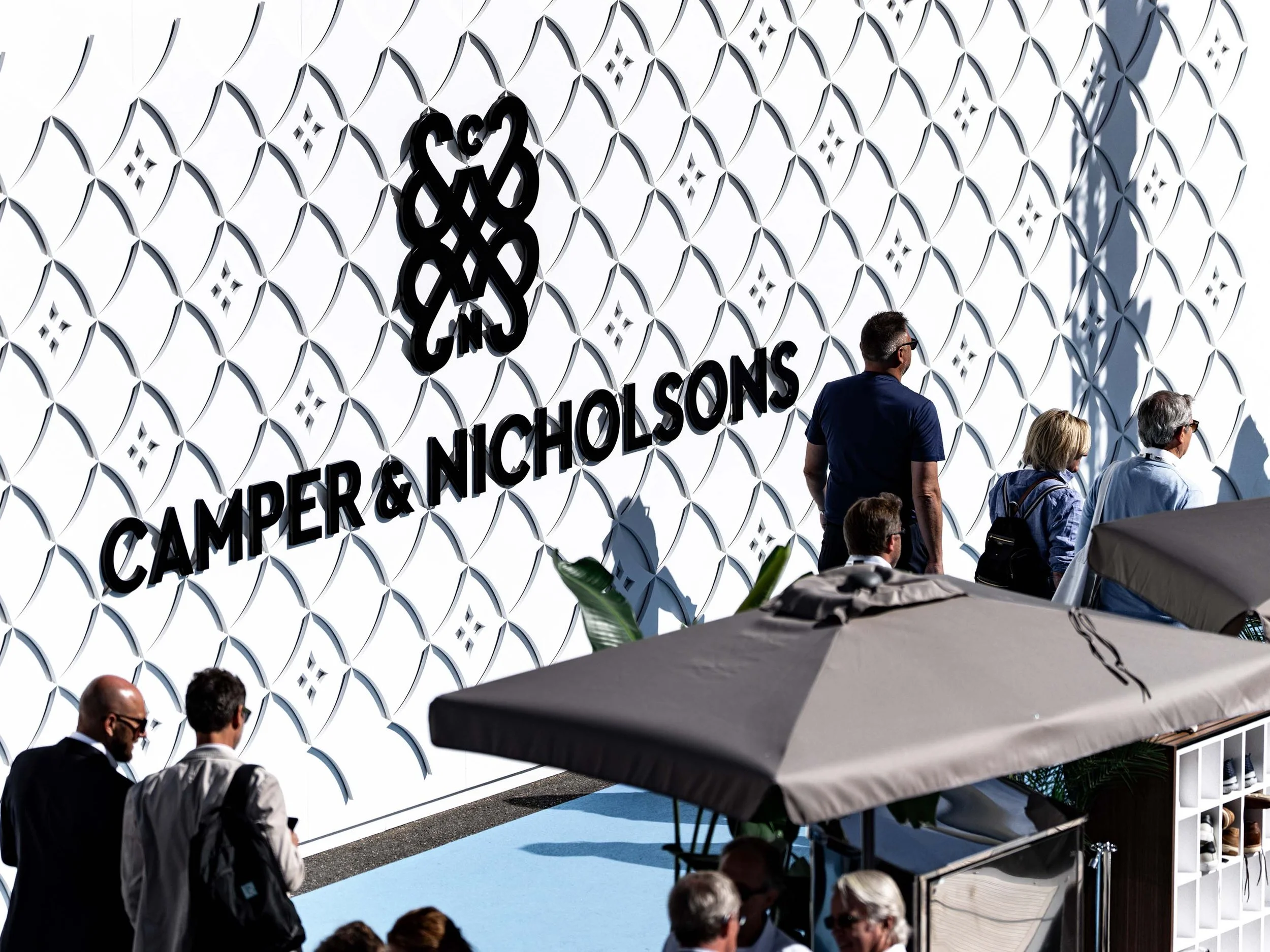 Image of the C&N stand at a yacht show. The wall of the stand shows the pattern structure in white and the emblem and logotype in black. People walk around