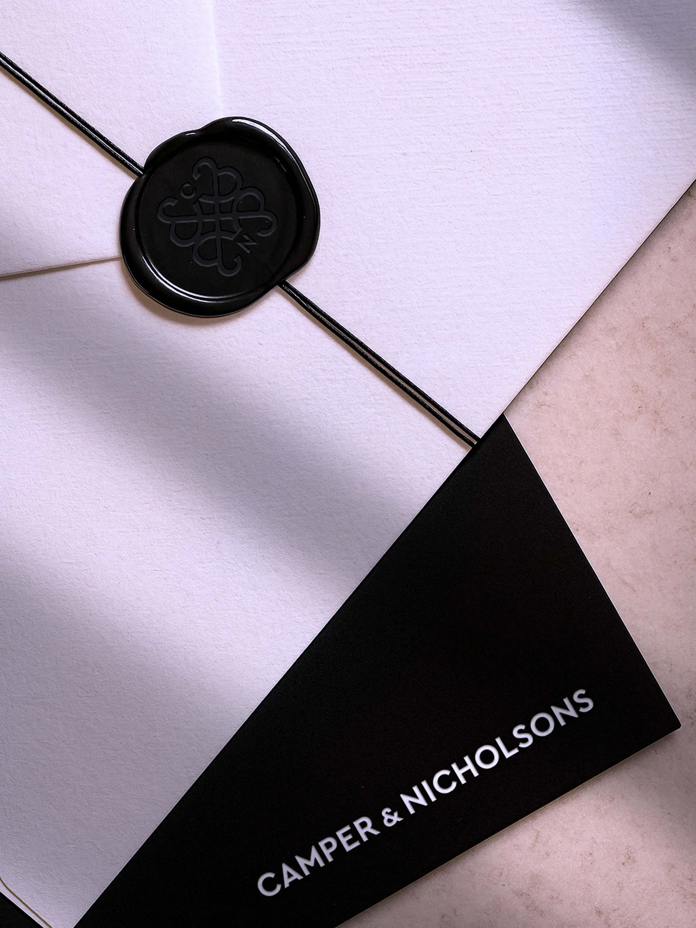 A close up image of a white envelope and a black card. The envelope has a black wax seal with the Camper & Nicholsons emblem. The black card has the logotype embossed in white foil