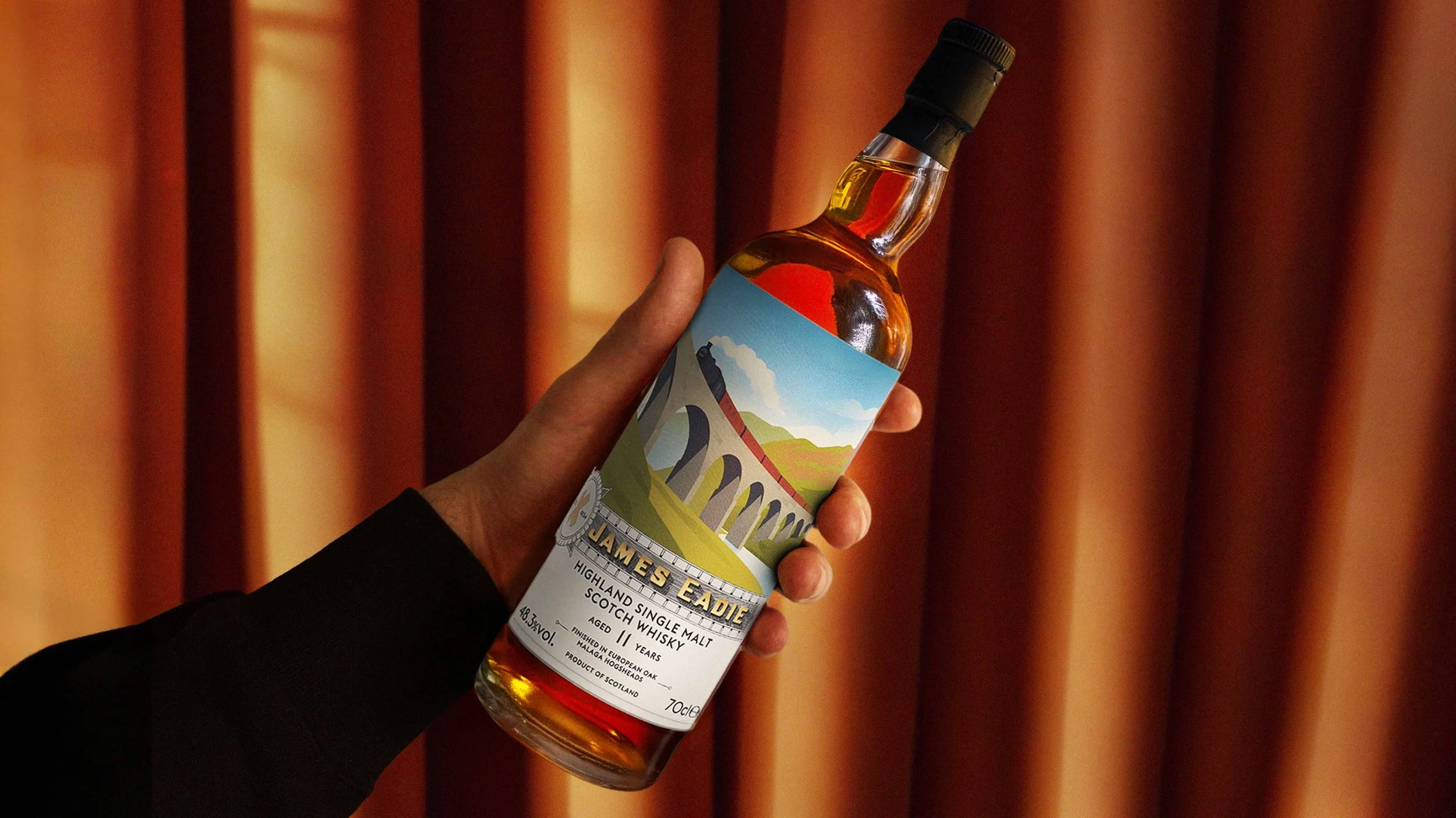 A hand holding the Highland bottle. The background is an orange thick curtain