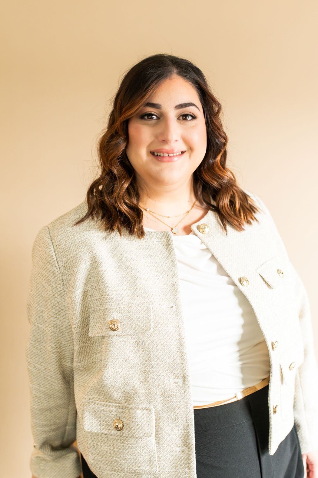 Woman with wavy brown hair wearing a cream jacket and black pants, smiling against a beige background.