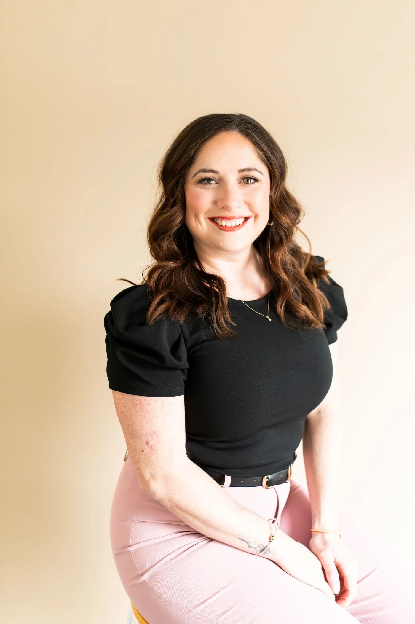 Smiling woman in a black top and pink pants sitting against a beige background.