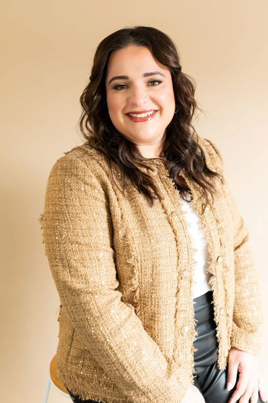 A woman with dark wavy hair smiling, wearing a gold textured blazer over a white top, sitting against a beige background.