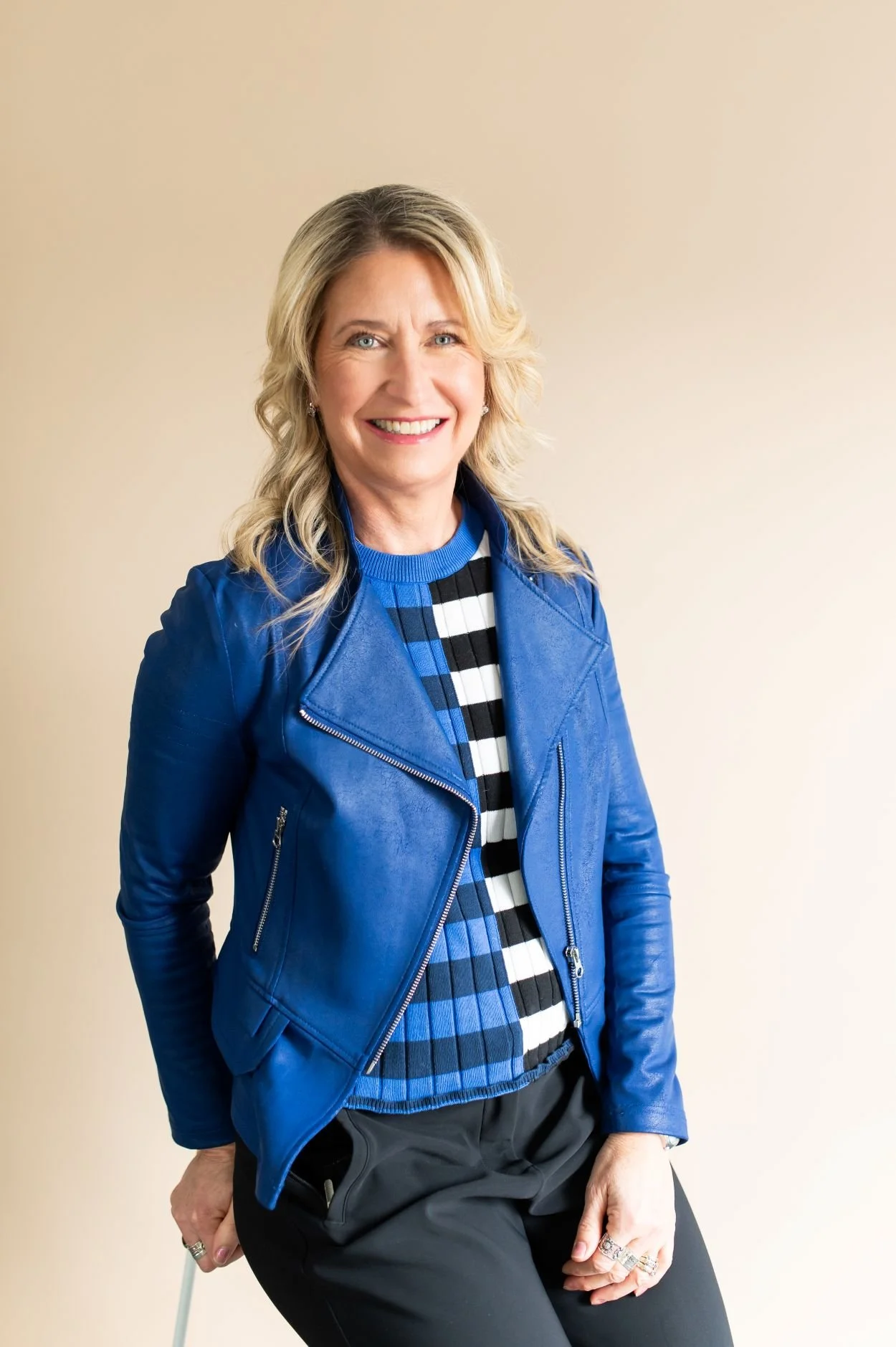 A female business owner with blonde wavy hair, smiling, wearing a blue leather jacket, a striped blue, black, and white top, and black pants, sitting against a plain beige background.