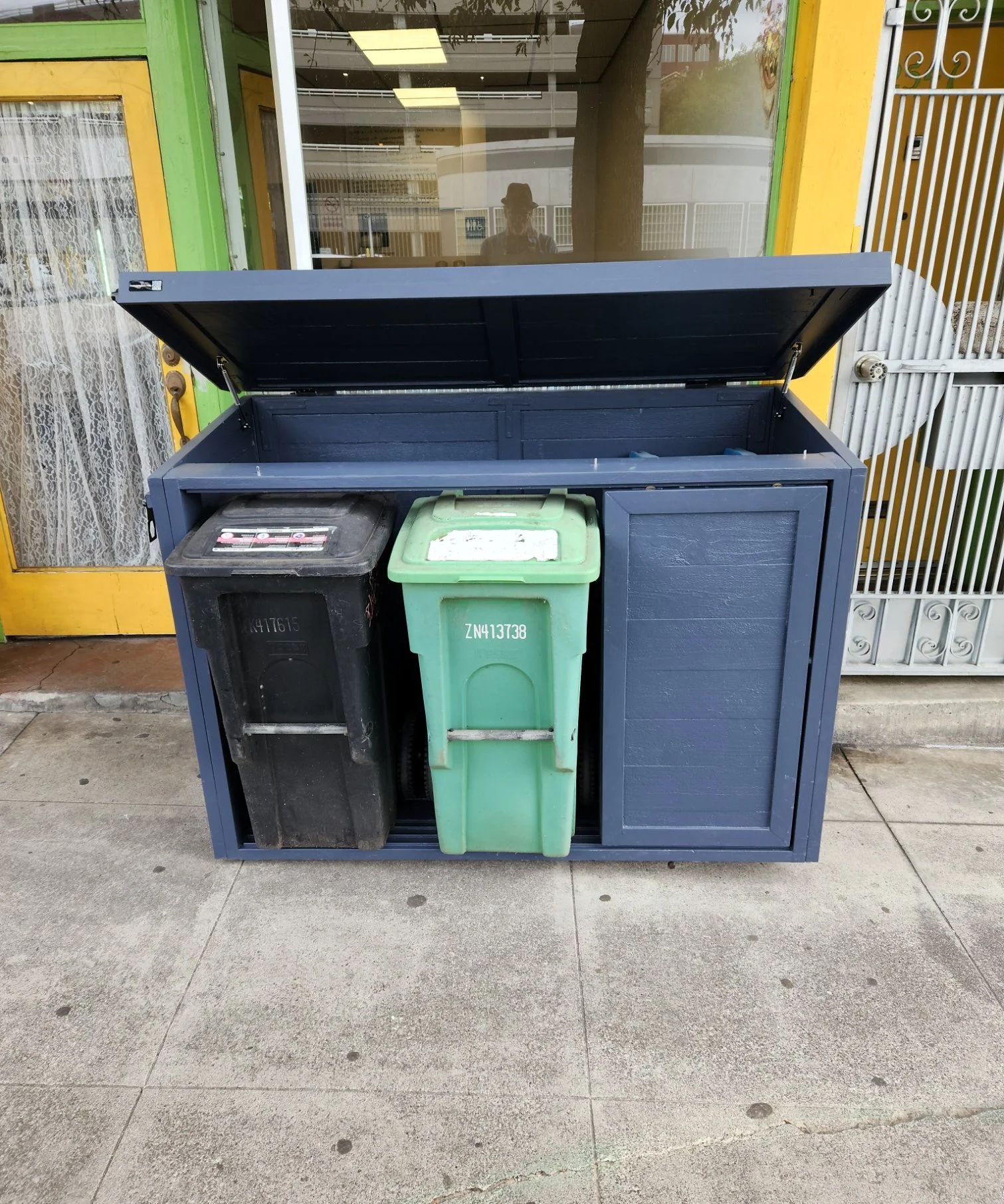 A black, green, and blue trash and recycling bin outside a building, with the lid open, showing no trash inside. The building has yellow and green walls, and a door with lace curtains. The ground is concrete.