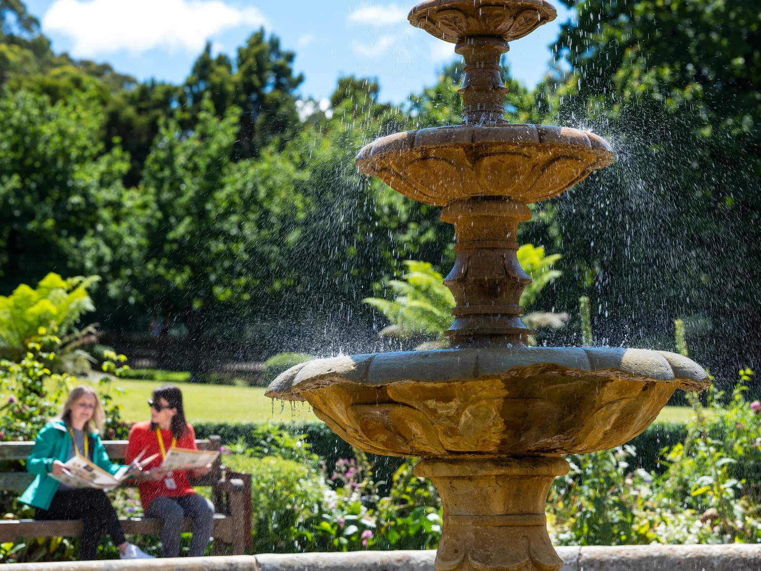 Outdoor fountain in a park with water spraying, two people sitting on a bench in the background, surrounded by greenery and trees.