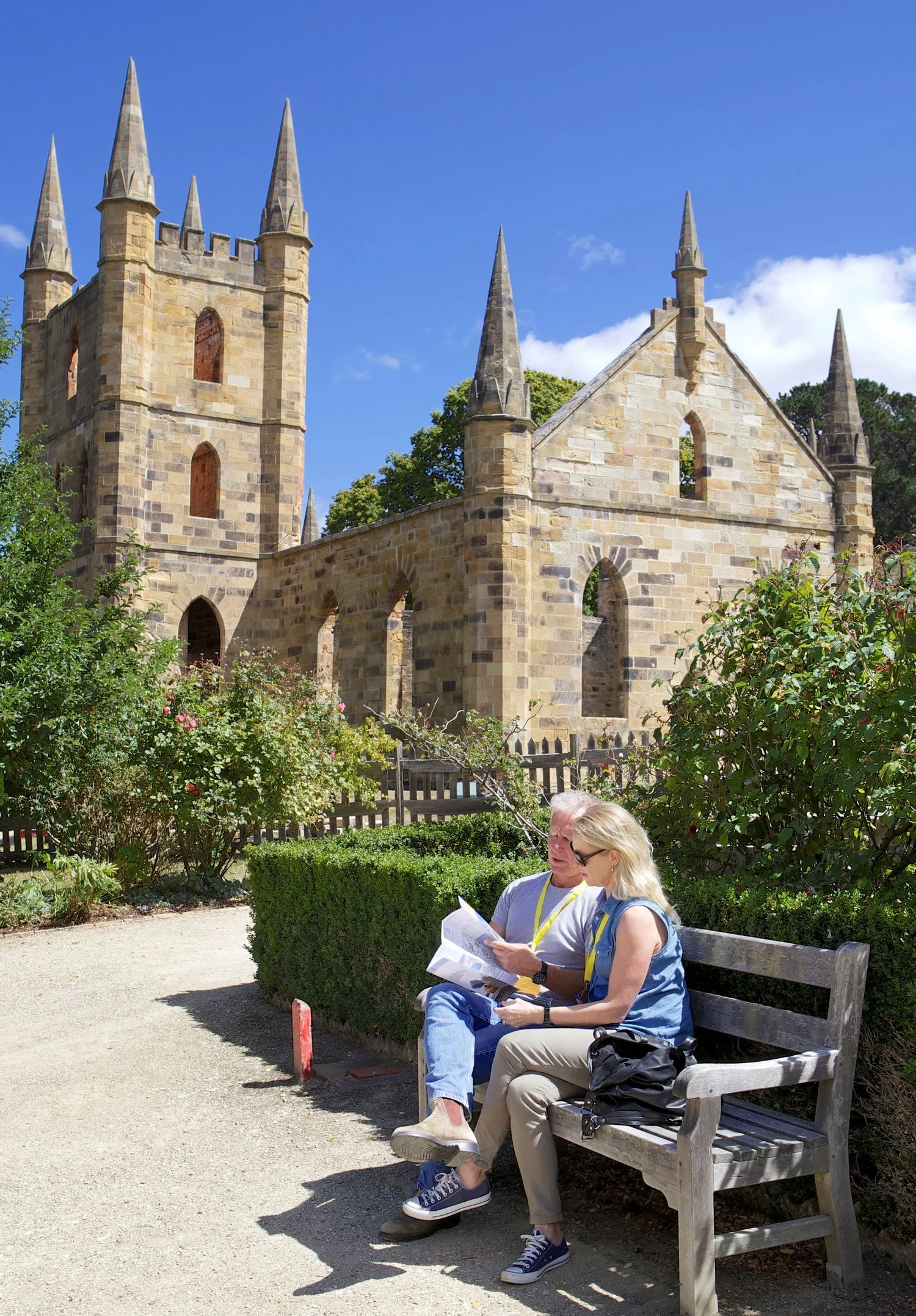 Historic church ruins with stone spires and arches. Two people sitting on a bench in the foreground, looking at a map or brochure. Surrounding garden with flowers and hedges. Clear blue sky.