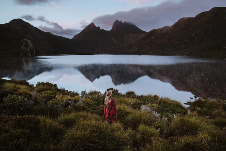 A person wearing a red plaid shirt and hat stands in front of a lake with mountain reflections at dusk, surrounded by grassy vegetation.