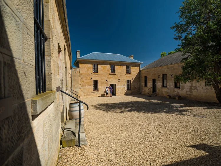 Historic stone buildings with blue sky, courtyard with gravel, tree, and wooden barrel beside a staircase.