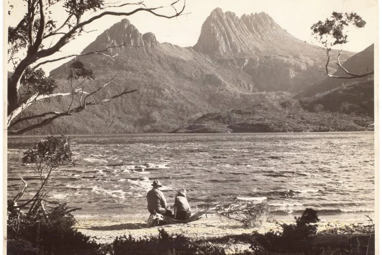 Two people sitting by a lake with mountains in the background, black and white photo.