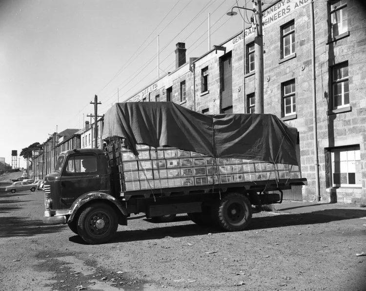 Vintage black and white photo of a truck with a covered cargo, parked on a street, with industrial buildings in the background.