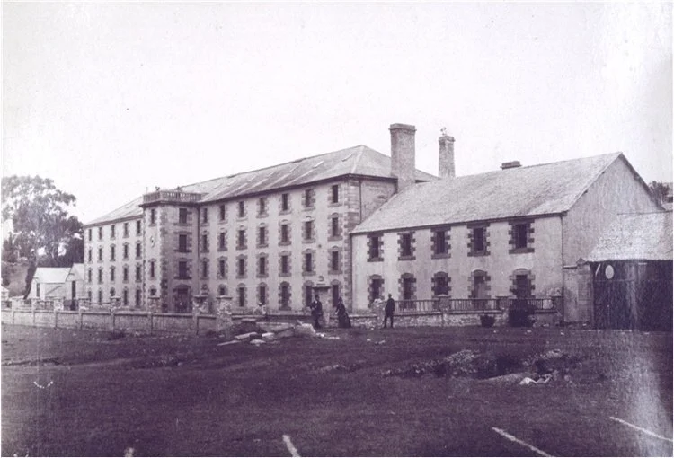 Historic stone building, likely from the 19th century, with multiple chimneys and a fenced courtyard, surrounded by grass and trees.