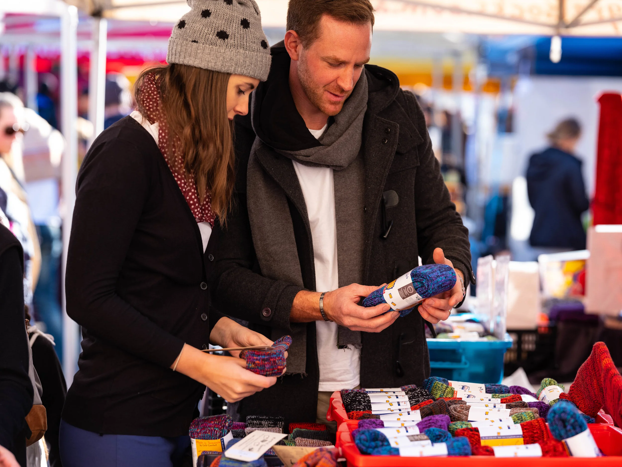 Two people shopping for knitted items at an outdoor market, examining colorful socks in a stall.