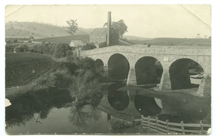 Historic stone arch bridge over a river with rural landscape in the background, including a building with a tall chimney and hills in the distance.