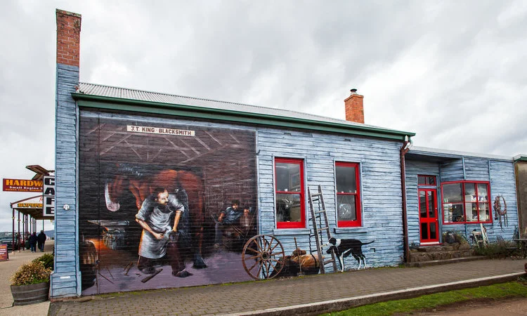 A mural of a blacksmith workshop on the side of a blue building with red window frames. The mural features a blacksmith working with a horse inside a barn. A sign reads 'J.T. King Blacksmith'. A cat and dog are painted near a wheel and ladder beside