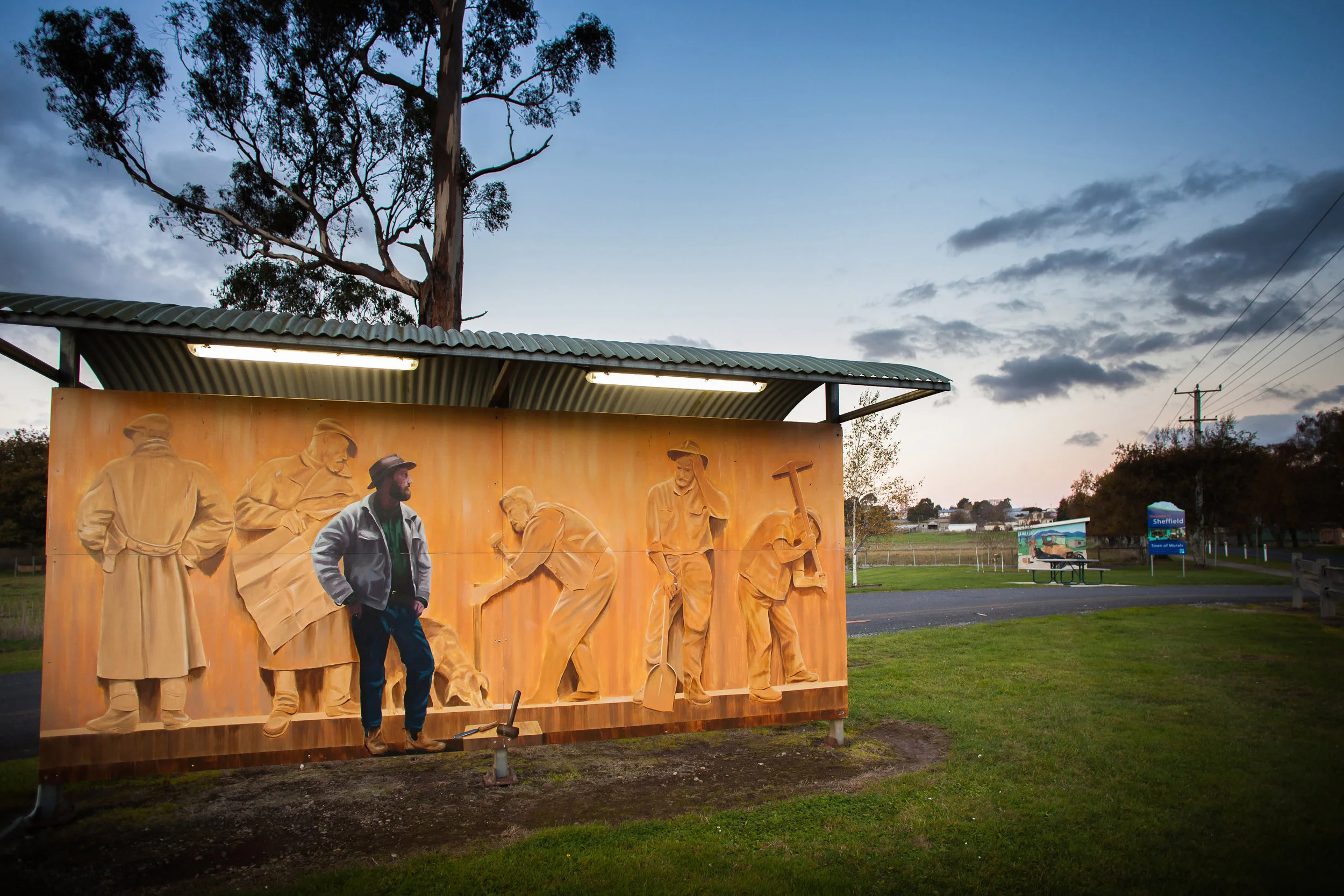Mural depicting historical miners in an outdoor pavilion with trees and grassy area in the background.