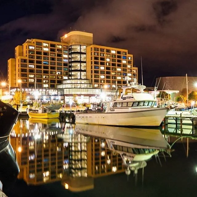 Night view of a harbor with boats docked, reflecting on calm water, against a backdrop of a lit-up building and cloudy sky.