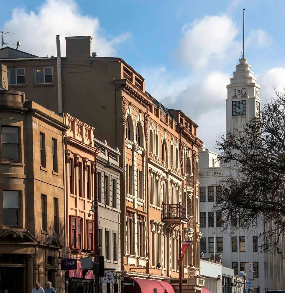 Street view with historic buildings, including a clock tower marked 'T & G,' under a blue sky with clouds.