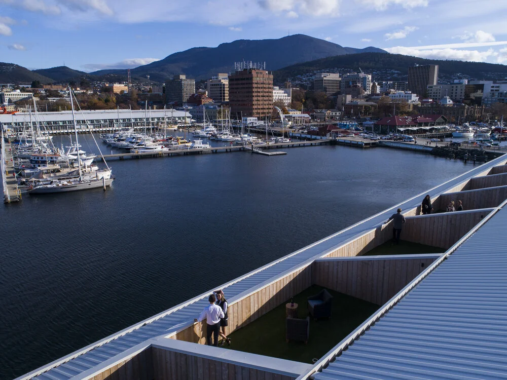 View of a harbor with boats docked, surrounded by city buildings and mountains in the background. People are standing on a rooftop with geometric design overlooking the scene under a partly cloudy sky.