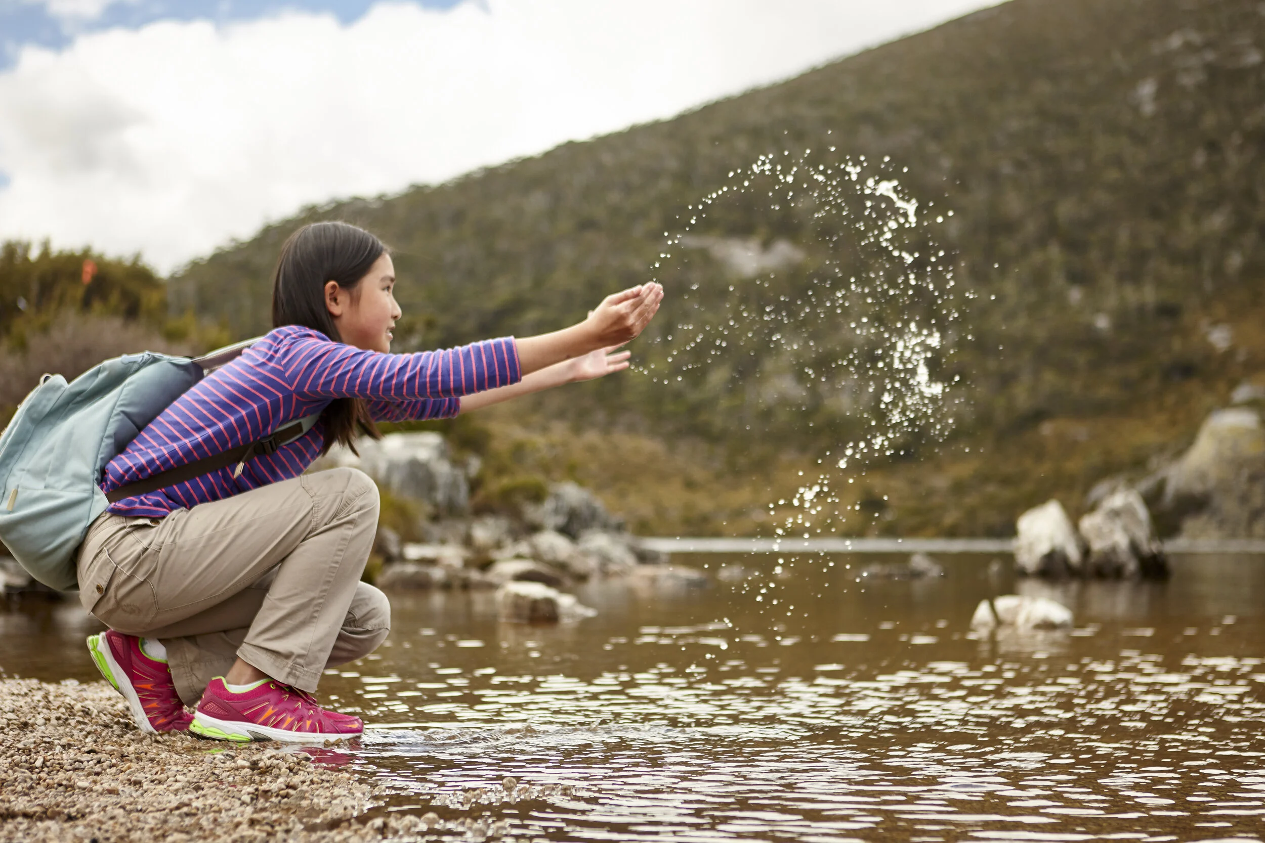 Girl with backpack playing by a lake, splashing water, with a mountainous landscape in the background.