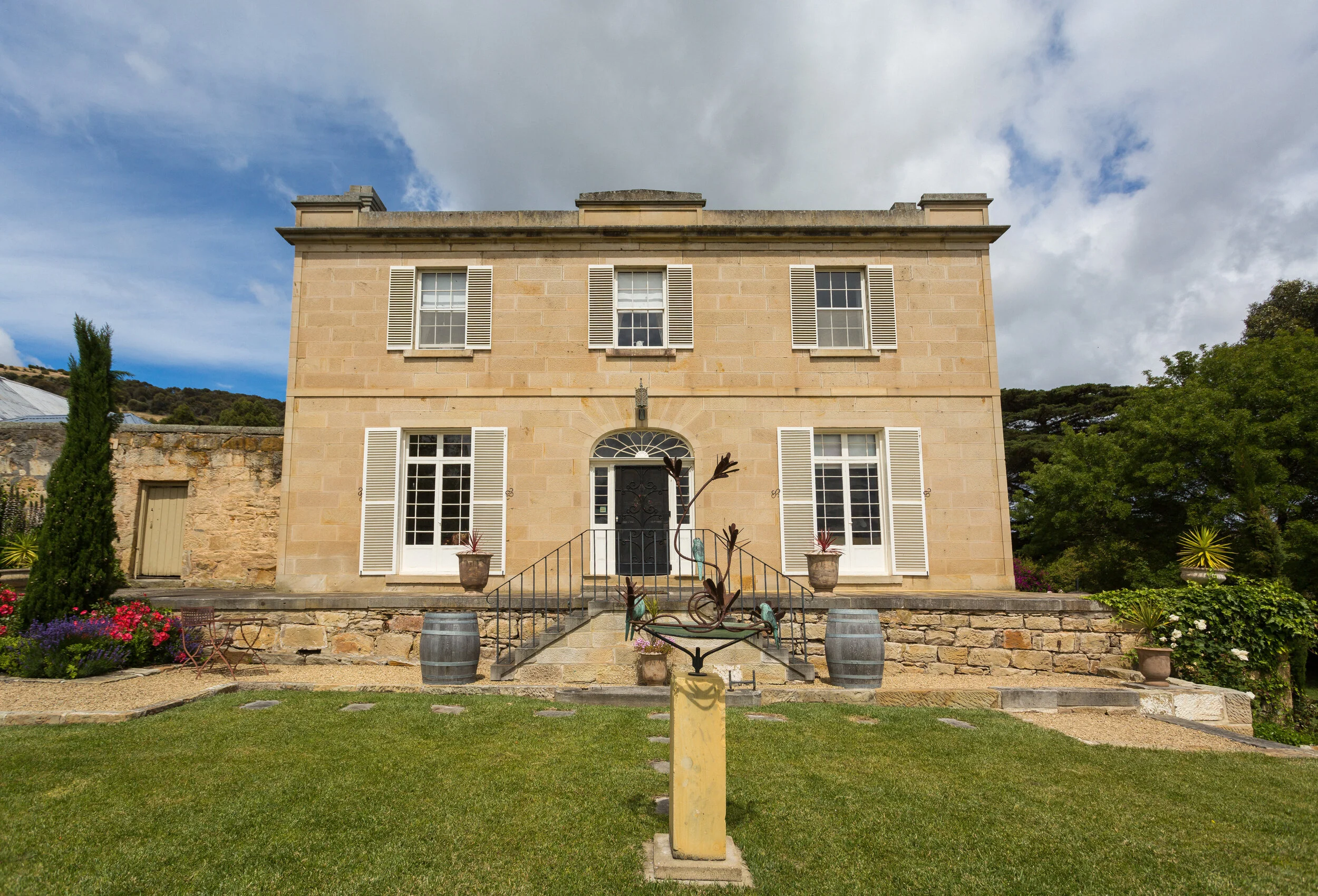 Historic two-story stone house with white shutters, surrounded by a well-maintained garden and lawn under a partly cloudy sky.