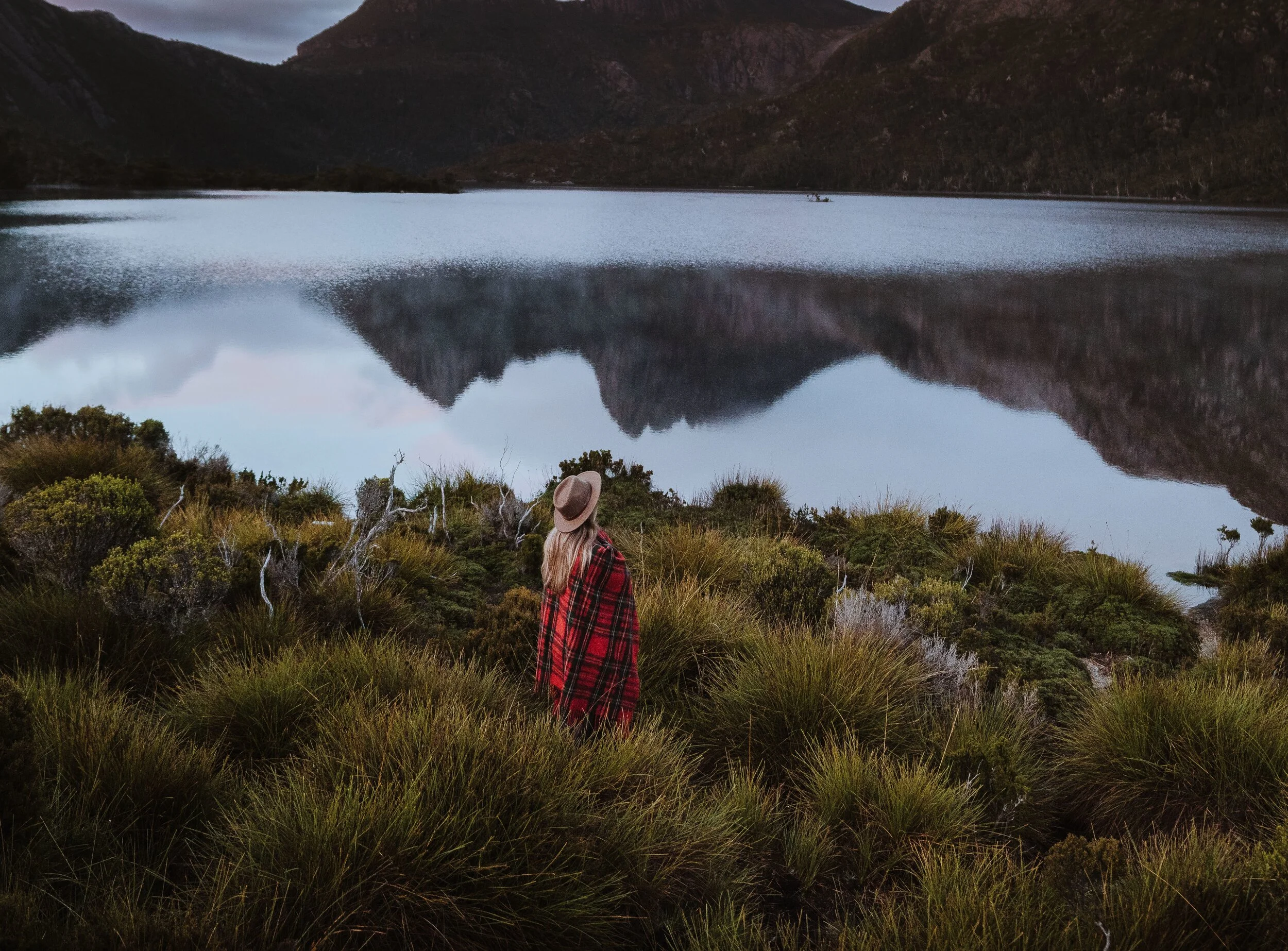 Person in red plaid jacket and hat stands near a lake with mountain reflection, surrounded by lush bushes and grass.