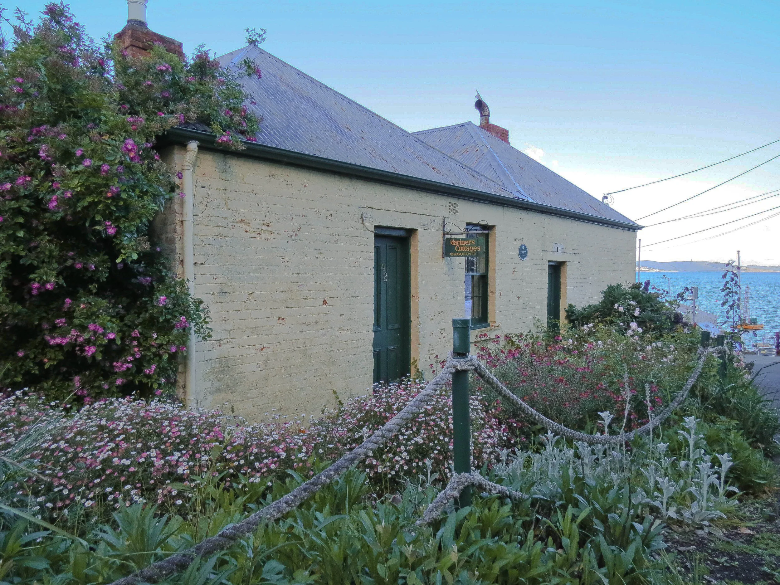 Historic cottage with a tin roof, surrounded by lush flower gardens, located near a waterfront.