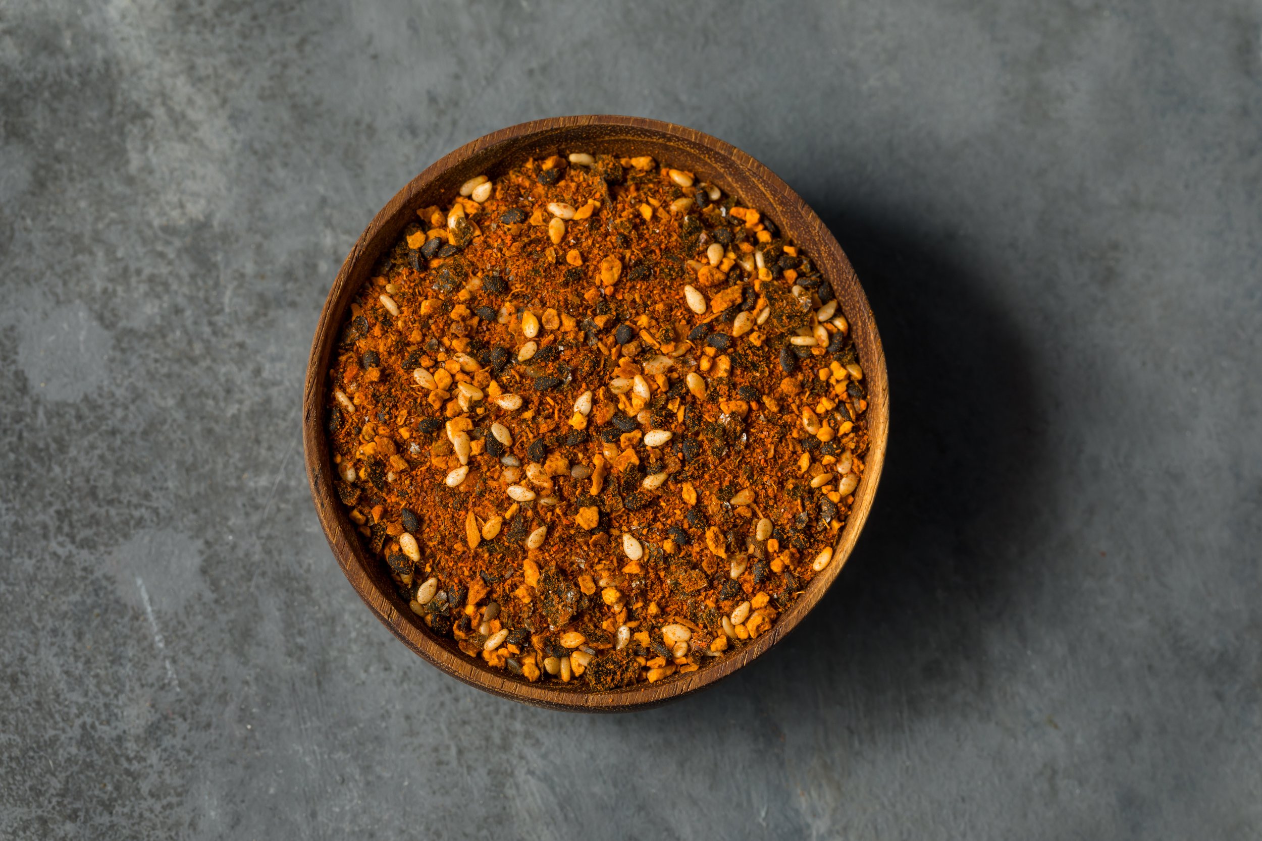 Close-up of a wooden bowl filled with a spicy seasoning blend containing red chili flakes, black pepper, sesame seeds, and other spices on a gray surface.