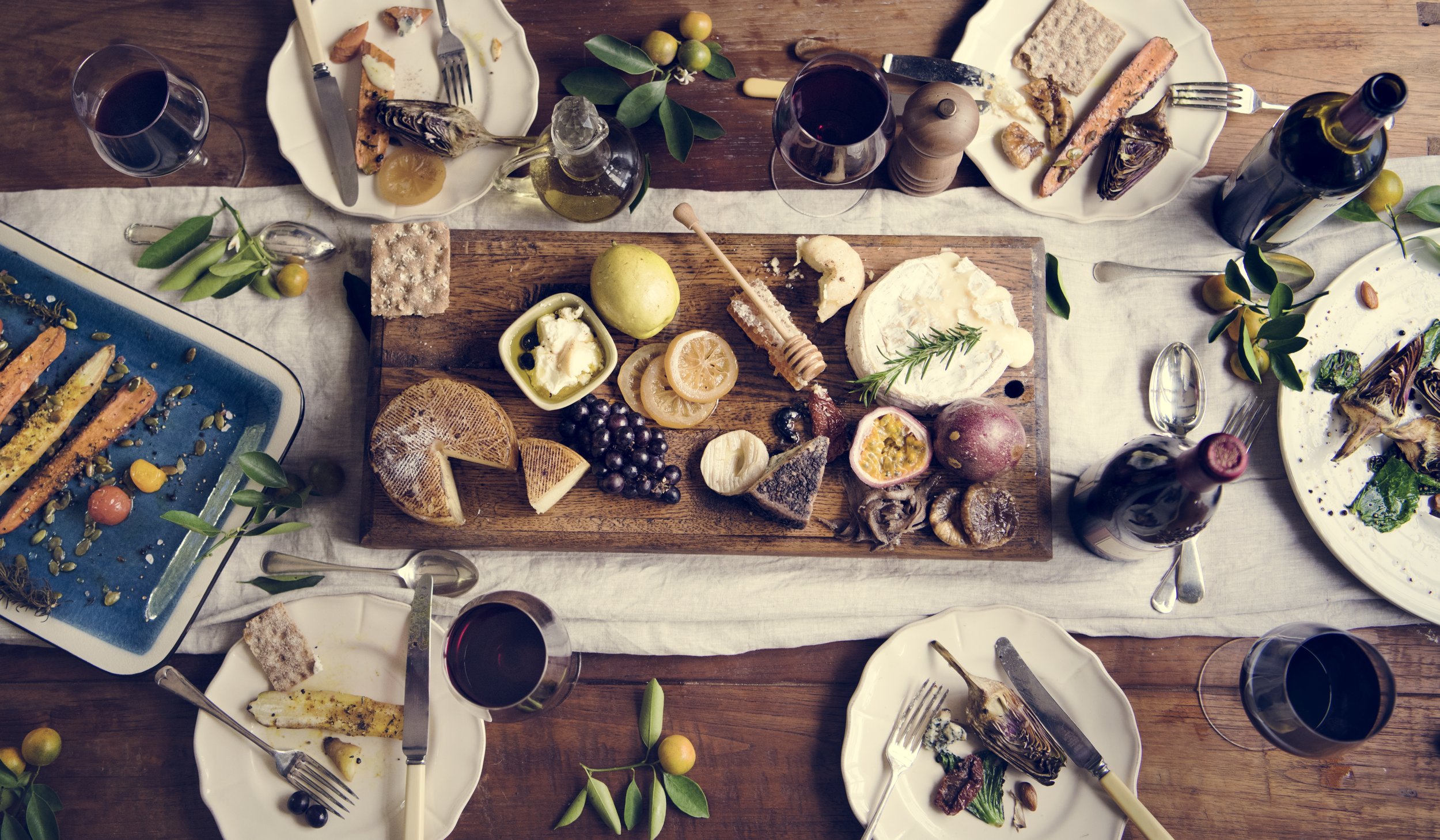 A top-down view of a large wooden dining table with various plates of food, glasses of red wine, and scattered green leaves and small fruits.