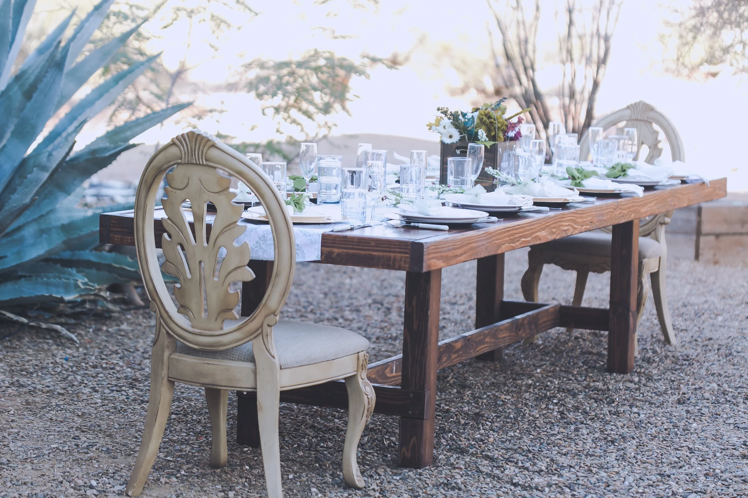Long wooden dining table with multiple empty glasses and white plates set for a meal. A central floral arrangement with white, purple, and green flowers. Elegant chairs with ornate backs surround the table. The setting appears outdoors with gravel gr