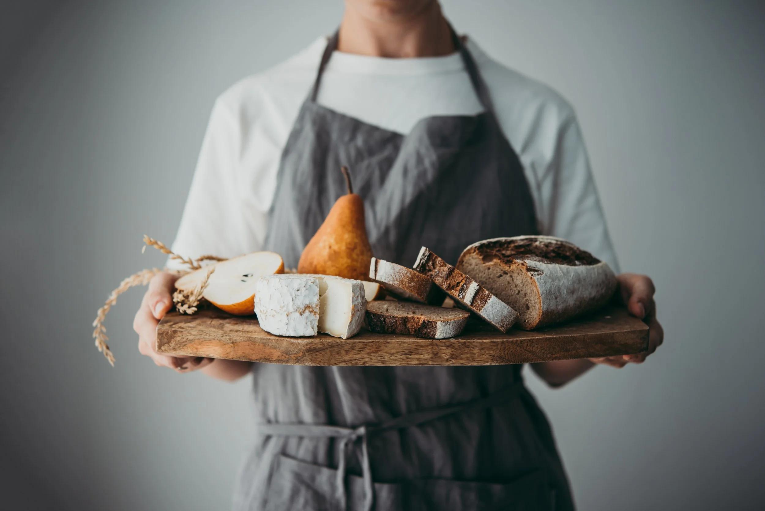Person holding a wooden cutting board with bread, cheese, pears, and sliced bread.
