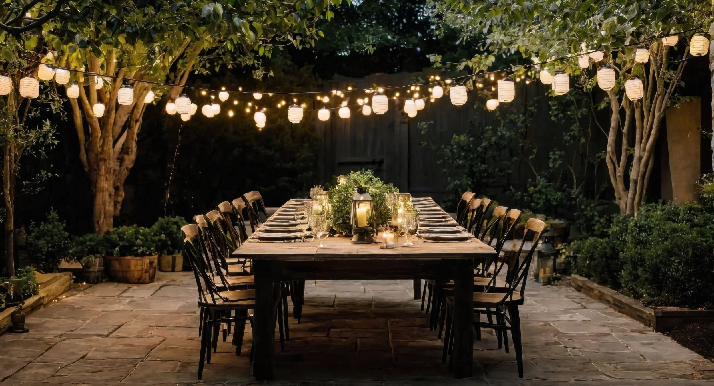 Outdoor dining table set for a gathering with candles and greenery, surrounded by trees and lit lanterns, at night.