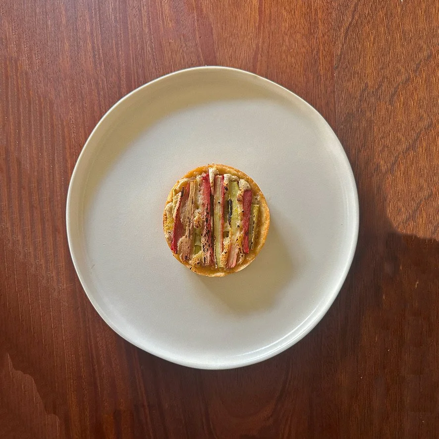 A small round savory pastry topped with slices of ham, pickles, and layers of other toppings, placed on a white plate on a wooden table.