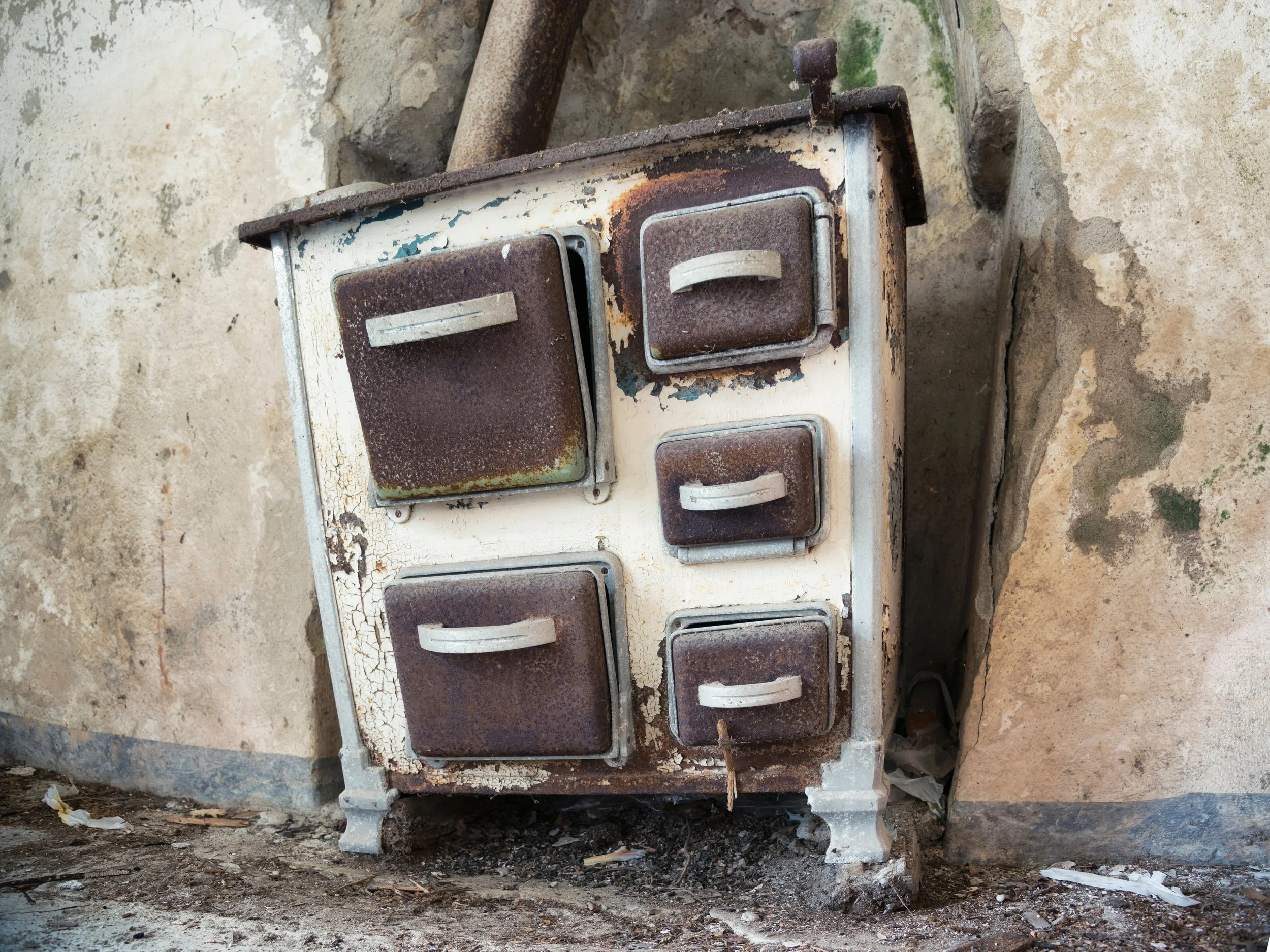 Old, rusty stove with four burners and a small oven, set against a peeling, stained wall with dirt and debris on the ground.
