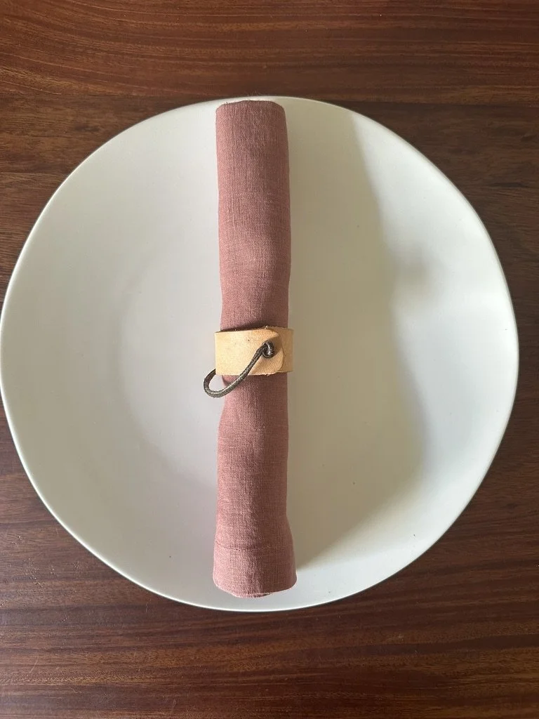 A white plate with a rolled pink cloth napkin secured with a small beige napkin ring that has a metal loop, placed on a wooden table.