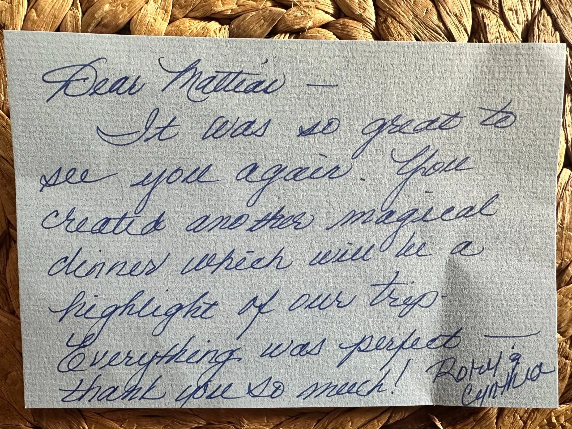 Close-up of a handwritten note on textured paper, with cursive writing in blue ink, placed on a woven surface.