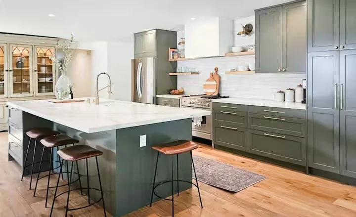 Modern kitchen with green cabinets, white countertops, and a large island with four stools. Includes stainless steel appliances, open shelves with dishes, and hardwood flooring.