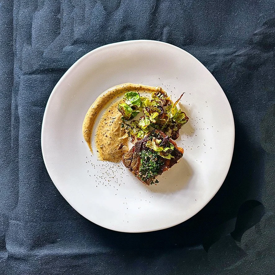 A white plate with a piece of grilled meat topped with greens, served with a side of mustard sauce, on a dark tablecloth.