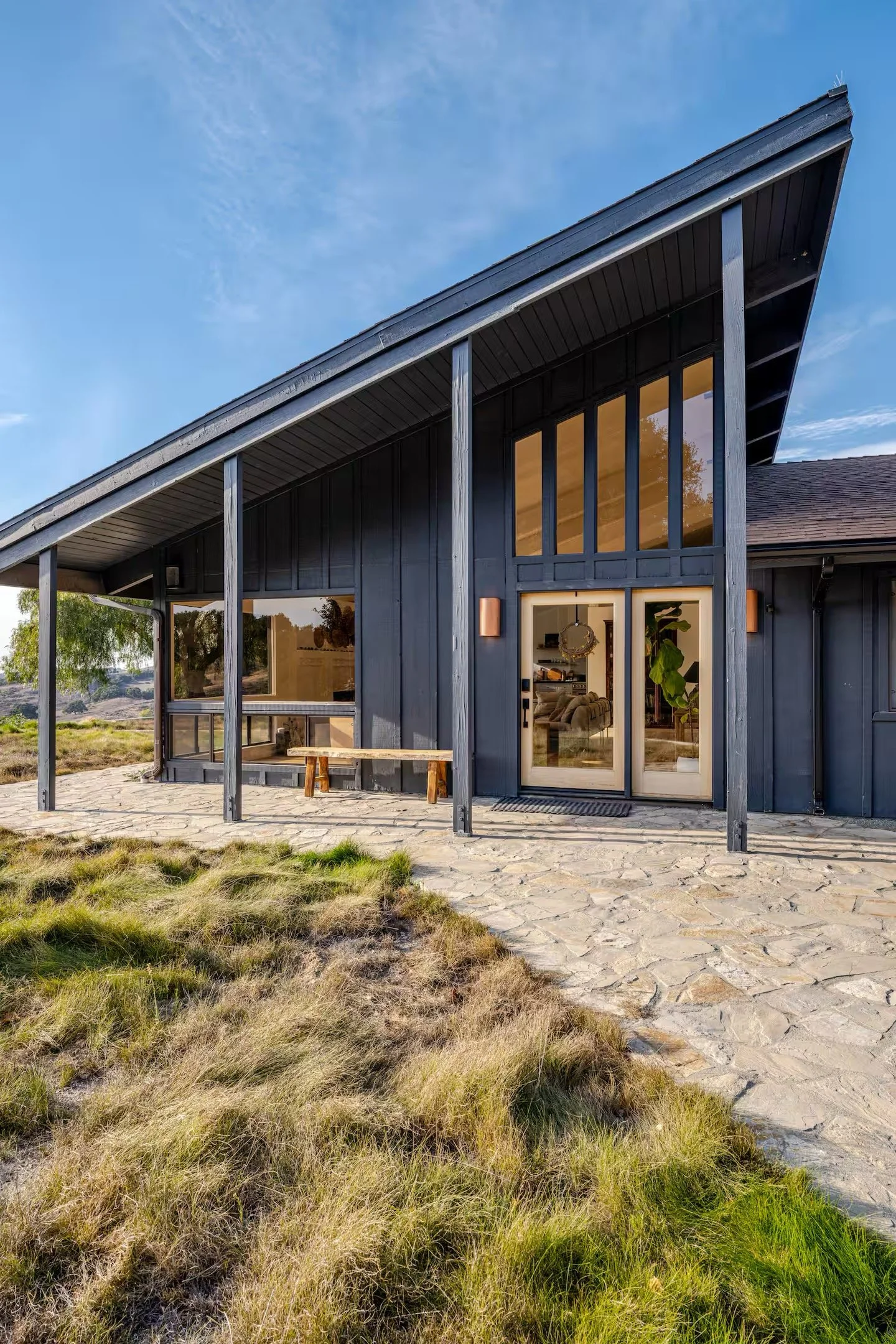 Modern house with black wooden siding, large glass doors, and a sloped roof, situated on rocky terrain with grassy patches.
