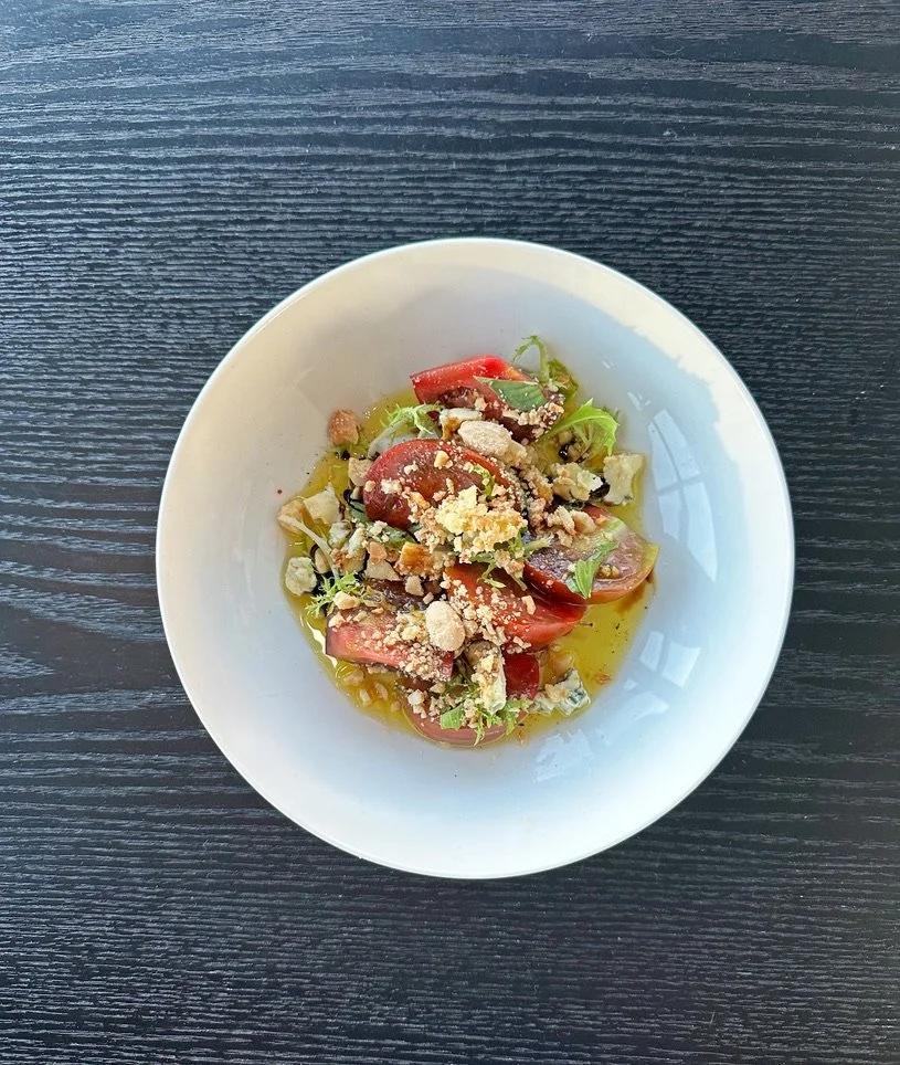 Salad with tomatoes, greens, crumbled cheese, and nuts in a white bowl on a dark wooden table.